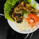 A grilled back pepper chicken leg on a bed of rice with lettuce and tomato slices. Two spice packets are in the background.