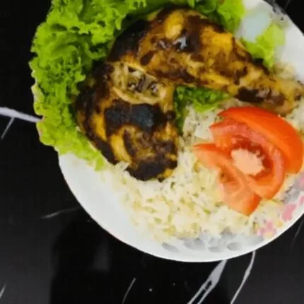 A grilled back pepper chicken leg on a bed of rice with lettuce and tomato slices. Two spice packets are in the background.