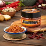 A bowl filled with vibrant chili powder next to a jar of spicy mama cripsy spicy anchoy on a wooden surface.