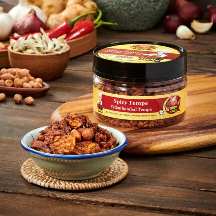 A jar of spicymama spicy tempe beside a bowl filled with roasted peanuts tempe on a wooden surface.