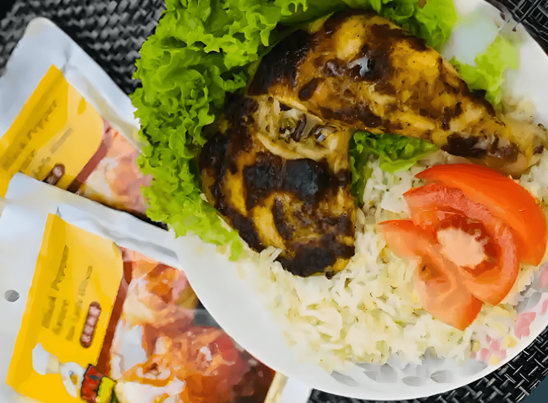 A grilled back pepper chicken leg on a bed of rice with lettuce and tomato slices. Two spice packets are in the background.