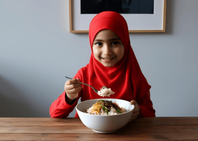 A young girl wearing a red hijab enjoys a plate of rice and vegetables, smiling as she takes a bite.