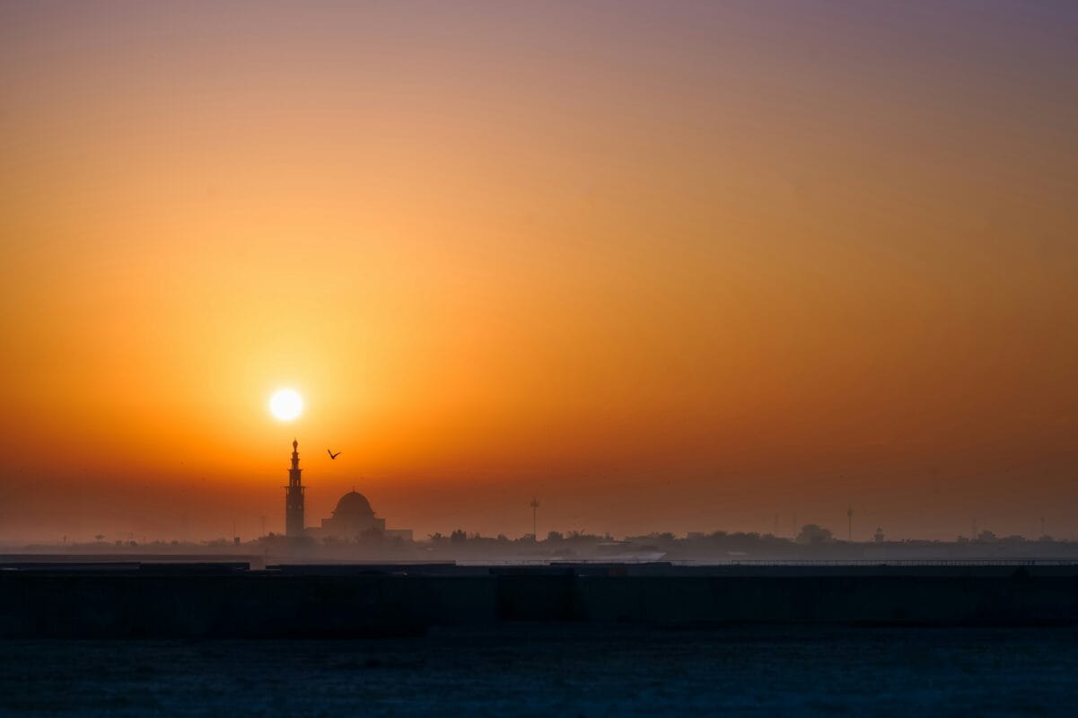 A sunset casts a warm glow behind a distant mosque, silhouetting its minarets against the colorful sky.