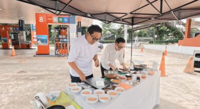 Two men cooking and arranging food at an outdoor food stand, surrounded by various ingredients and cooking equipment.