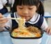 A young girl happily eating noodles at a school cafeteria table surrounded by classmates.