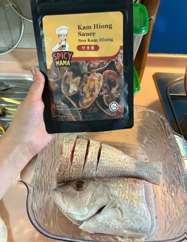 A person holding a packet of SpicyMama Kam Hong Hong sauce, showcasing its vibrant packaging and branding. with a plate of raw fish