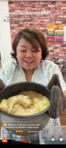  A woman displays a pot  of chicken rice in front of her, highlighting the meal with a smile.